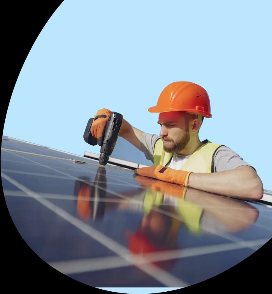 a man installing a solar panel on the roof of a house during the day. He wears a hard hat, gloves and tools as he carefully adjusts the panel with a drill. The sky is clear and cloudless, suggesting good conditions for work.